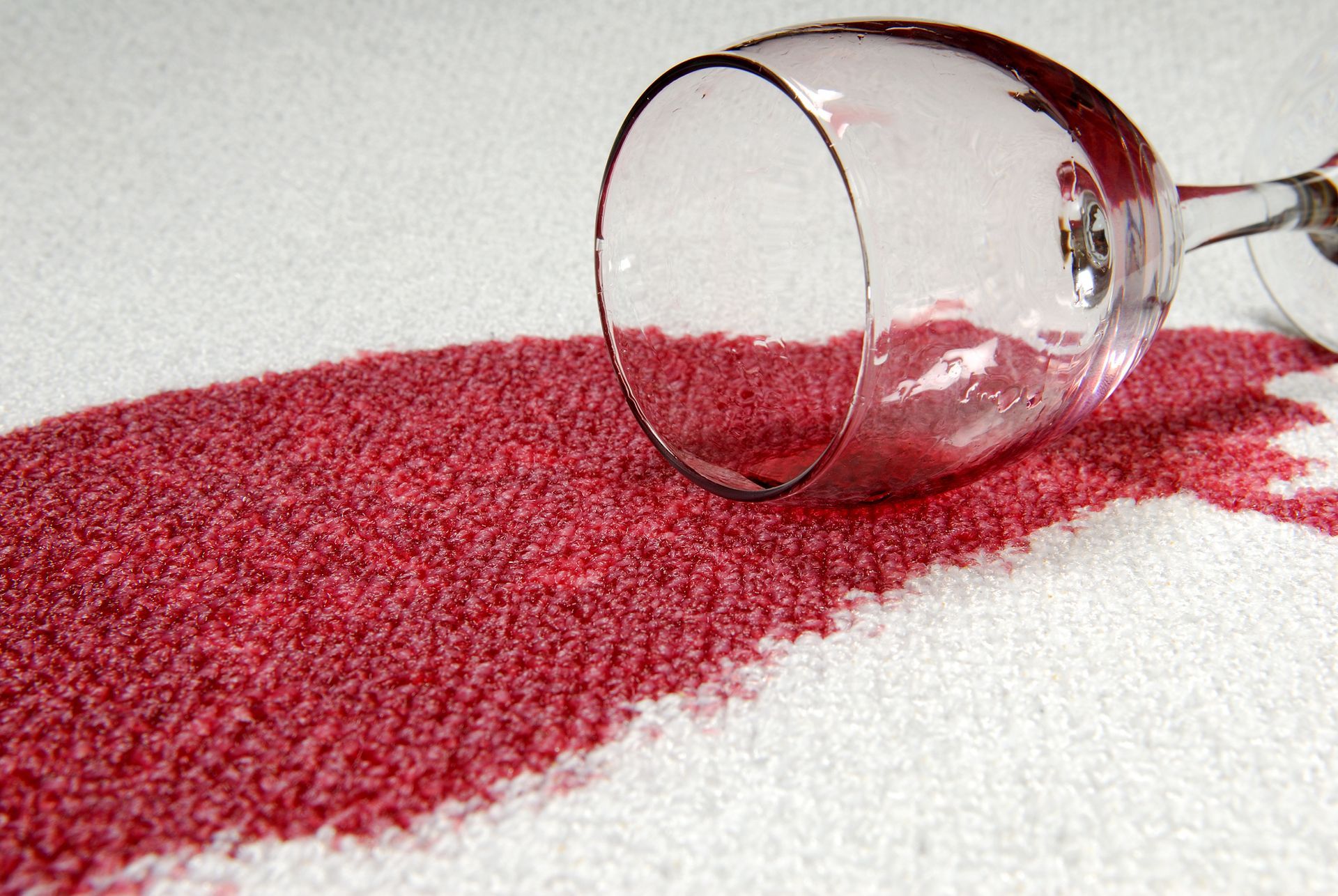 Wine glass tipped over, spilling red wine onto a white carpet.