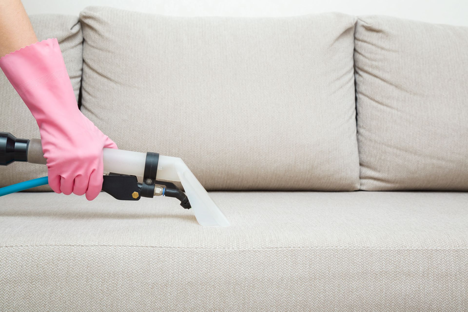 Person wearing pink gloves cleaning a beige couch with a cleaning tool.