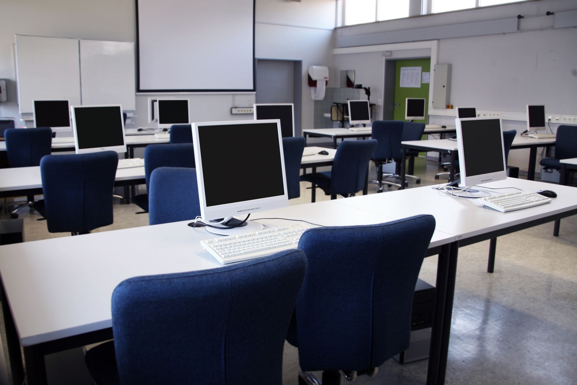 Computer lab with rows of white desks, blue chairs, and monitors. A projector screen hangs on the wall.