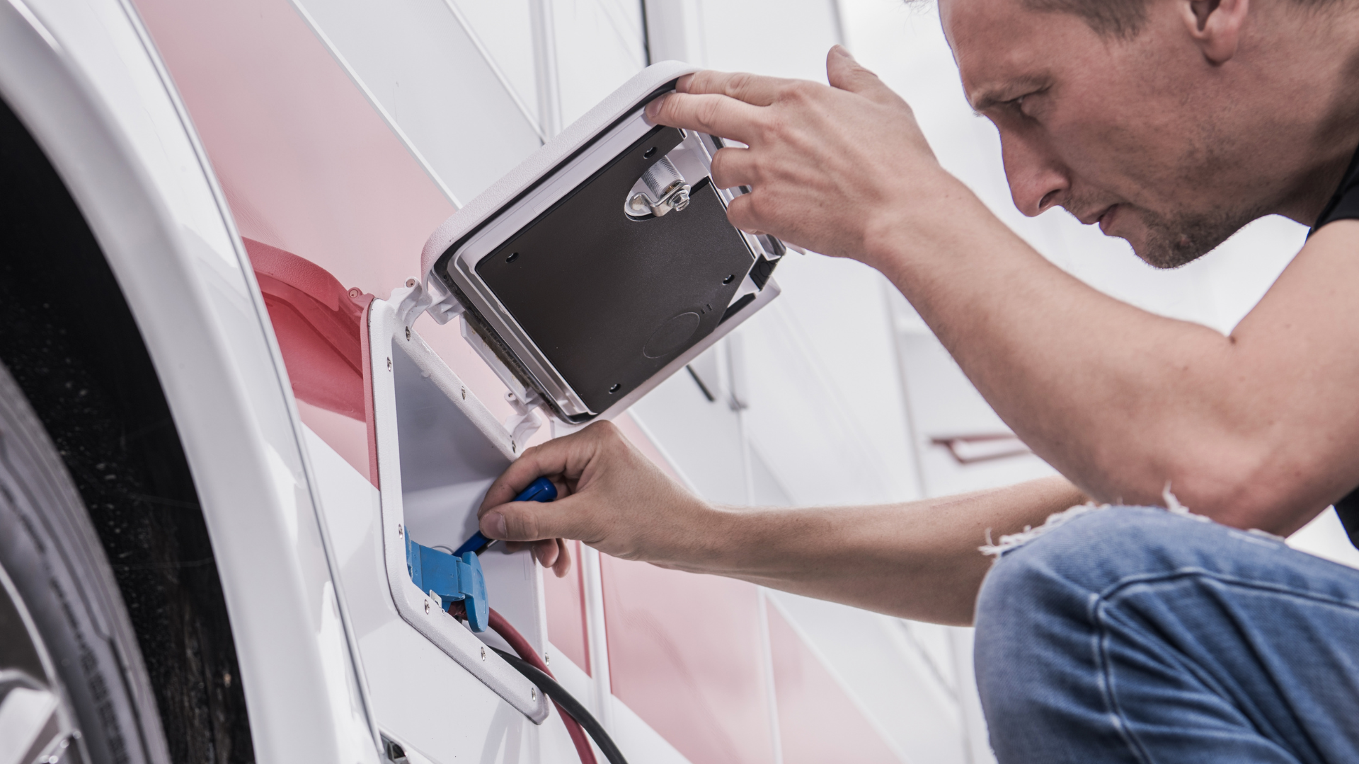 A man is kneeling down next to a camper and plugging a charger into it.
