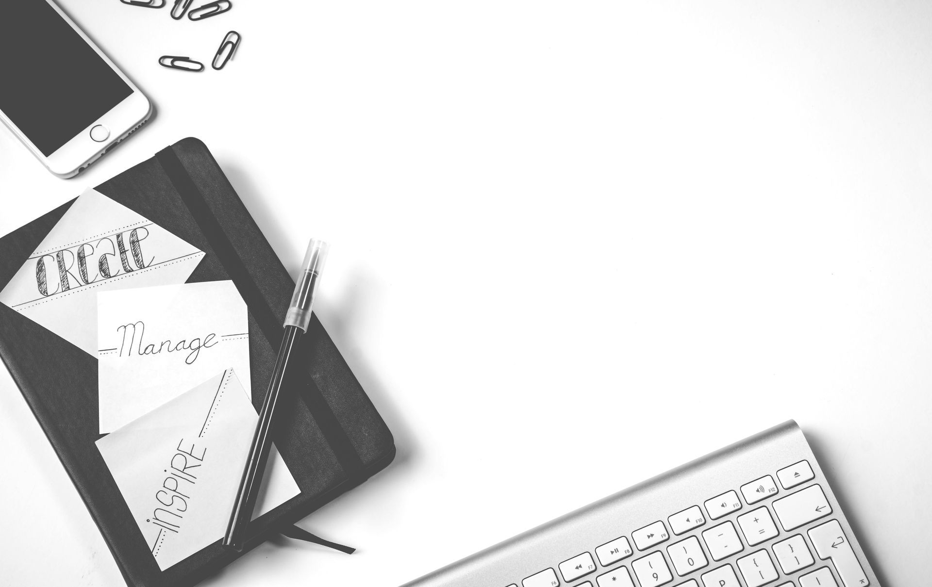 Desk scene: notebook, phone, keyboard, pen, paper clips on white surface.