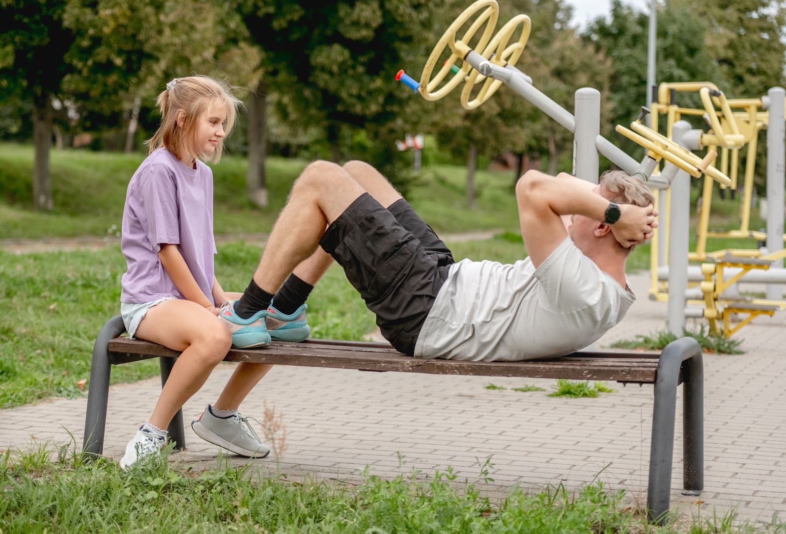 Two people exercising on outdoor gym equipment in a park: one lies on a bench, the other sits nearby.