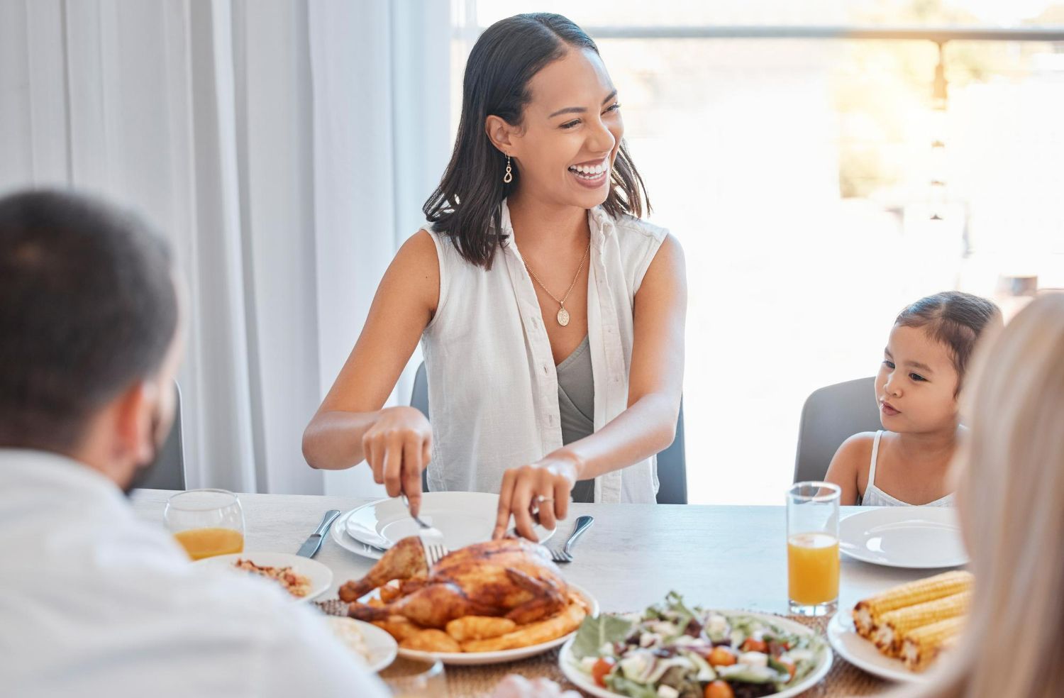 A family sits at a dinner table, smiling while one person carves a roasted chicken surrounded by side dishes.