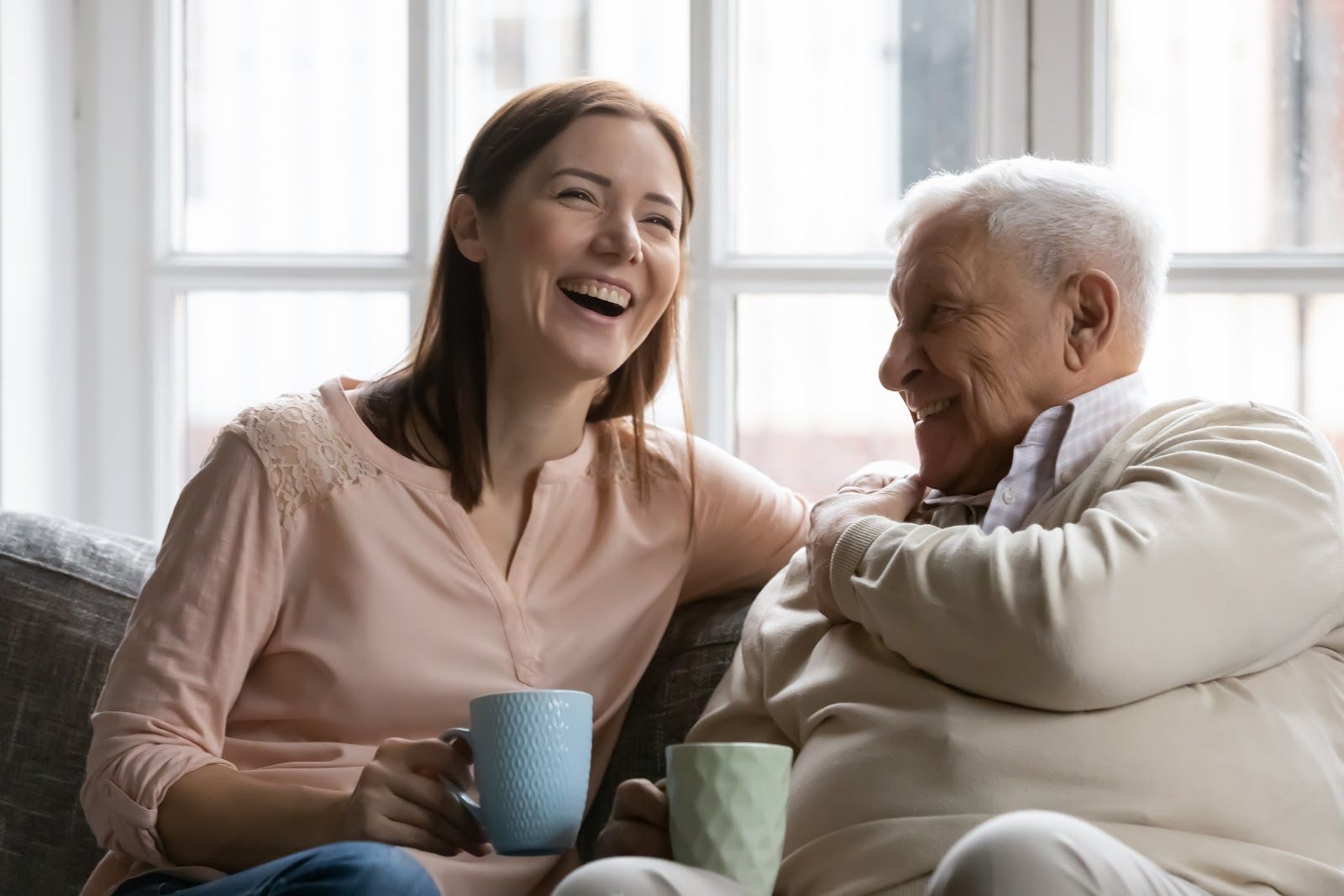 Two people smiling on a couch, holding mugs in a bright living room