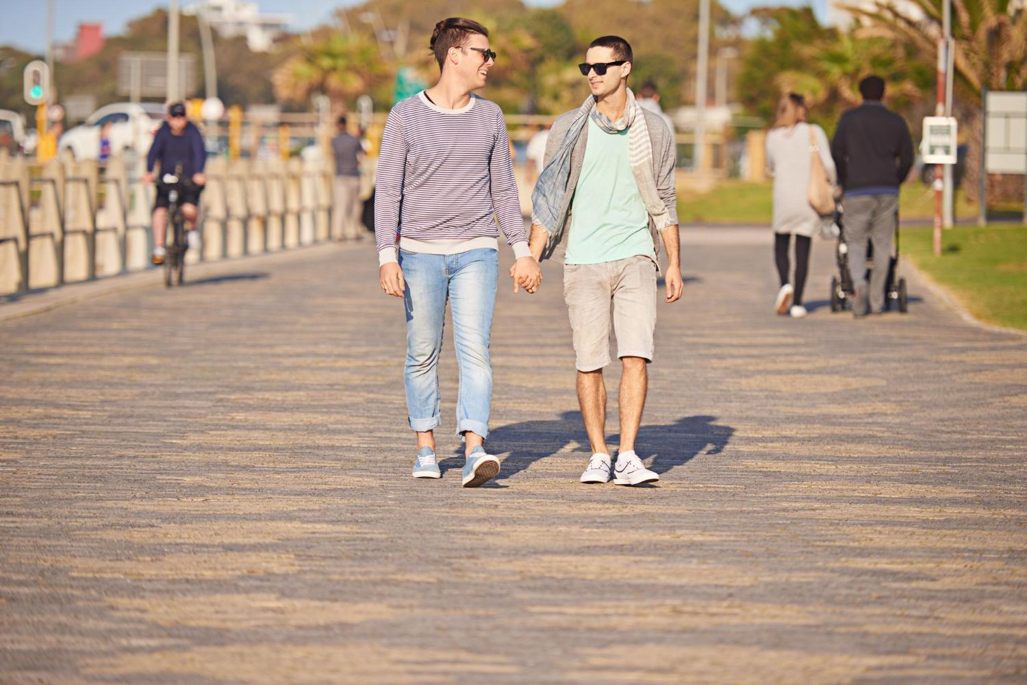 Two people walk hand-in-hand down a paved promenade on a sunny day, with others walking and cycling in the background.
