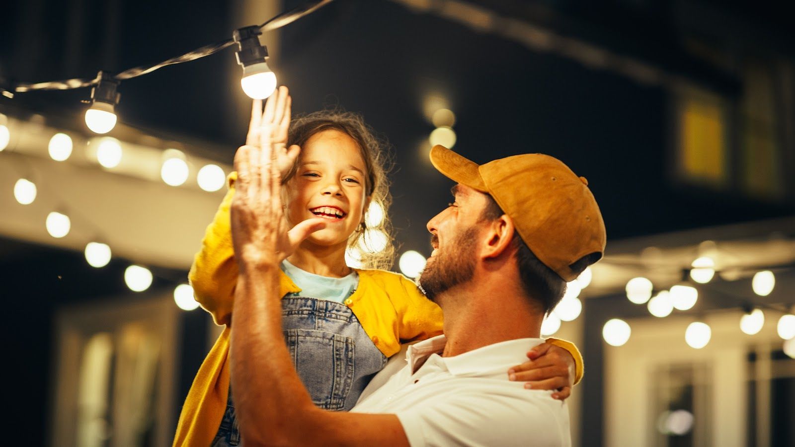 Adult lifting smiling child in a warmly lit outdoor setting with string lights