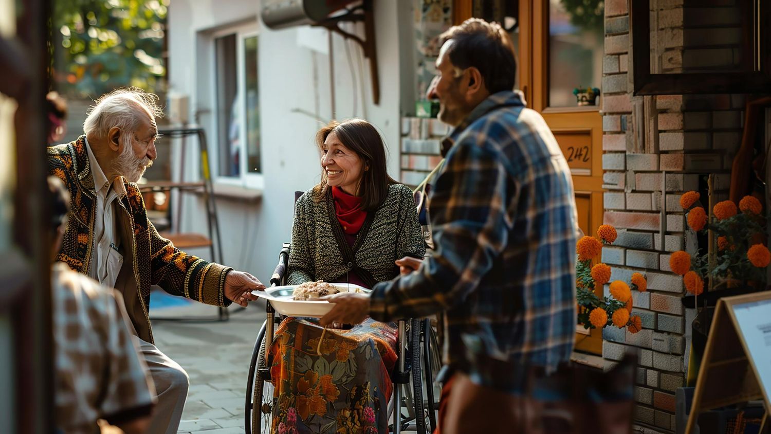 Three people interacting outdoors; one in a wheelchair holds a bowl while two others stand nearby in a casual setting.