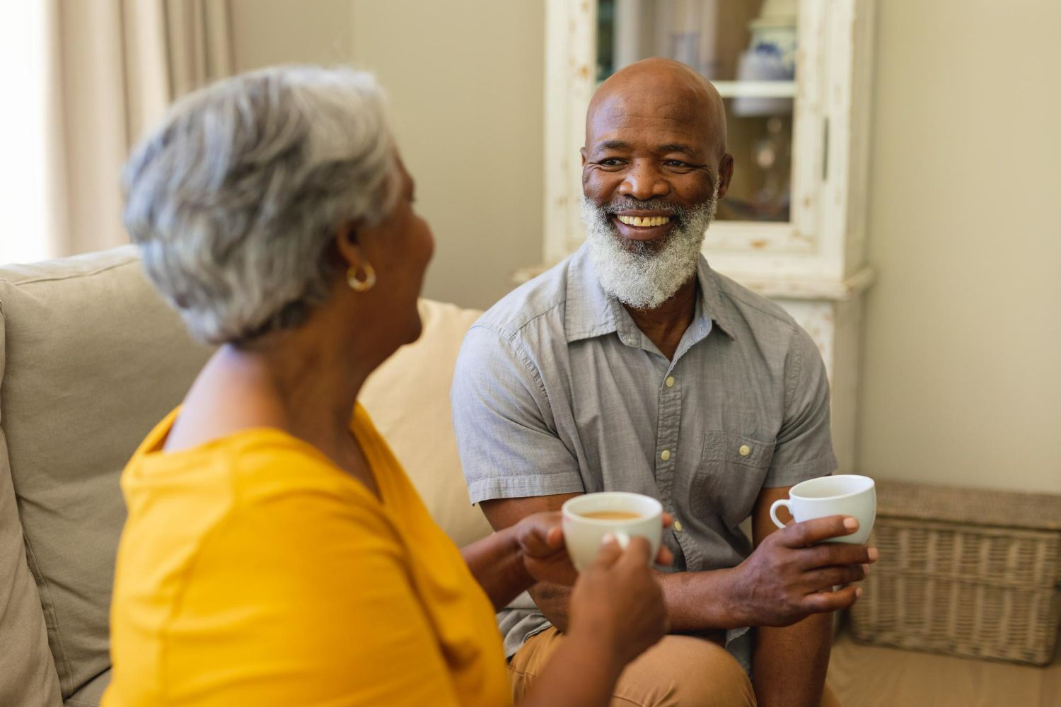 Two people sitting on a sofa, smiling and holding cups while engaged in conversation in a home setting.