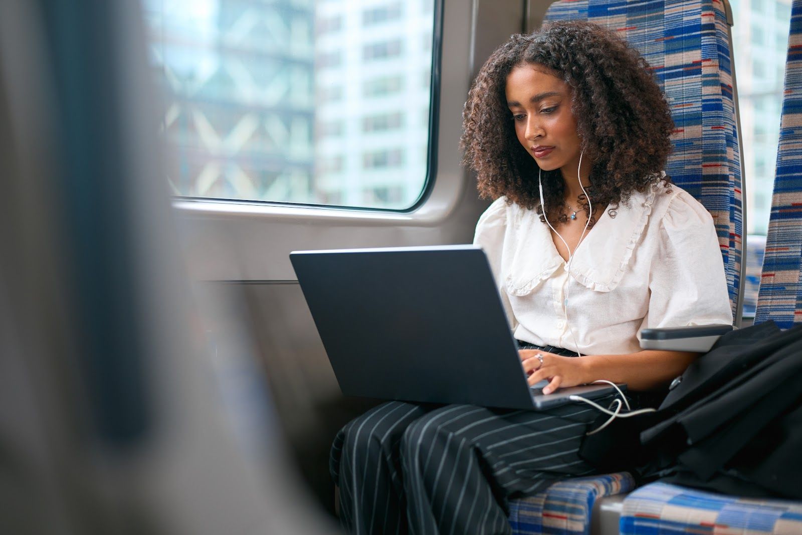Woman using a laptop on a train seat by the window, wearing a white blouse and striped pants.