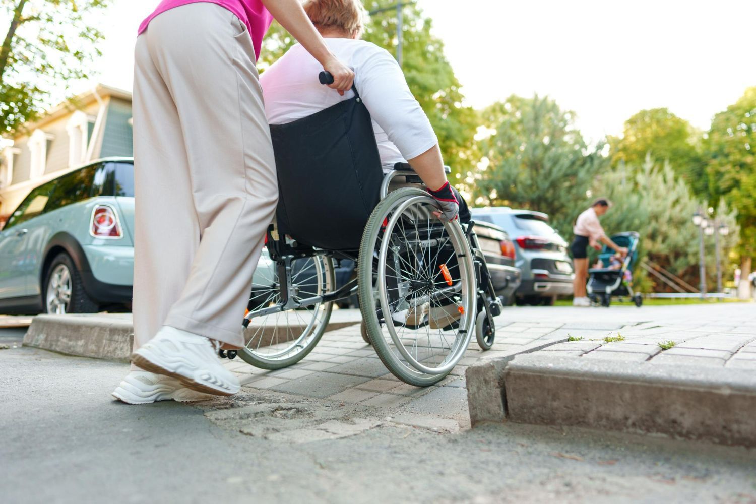 A caregiver pushes a person in a wheelchair along an outdoor paved path near a parked car.