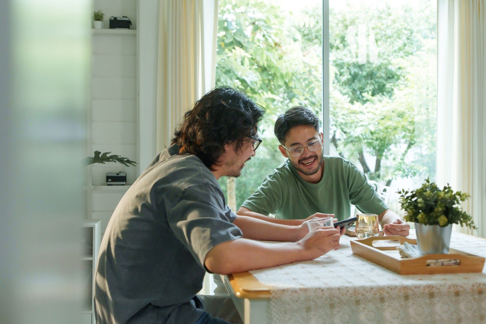 Two people chatting at a sunlit kitchen table with drinks and papers