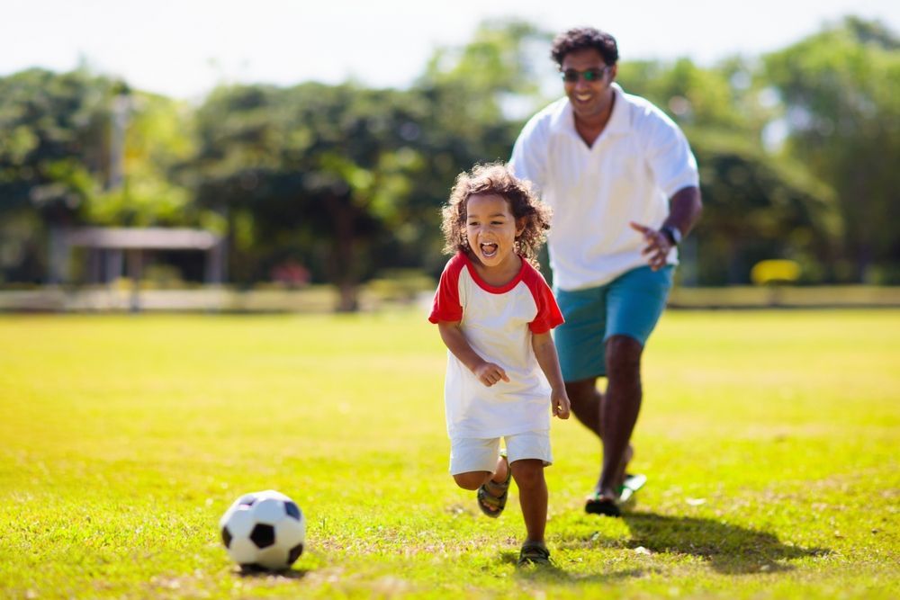 A child and an adult playing soccer on a grassy field, both smiling while running toward a ball.