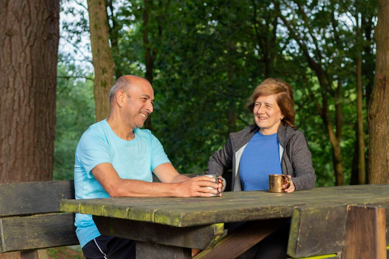 A person in a blue shirt and another in a grey jacket sit at a wooden picnic table in a forest, smiling and holding mugs.
