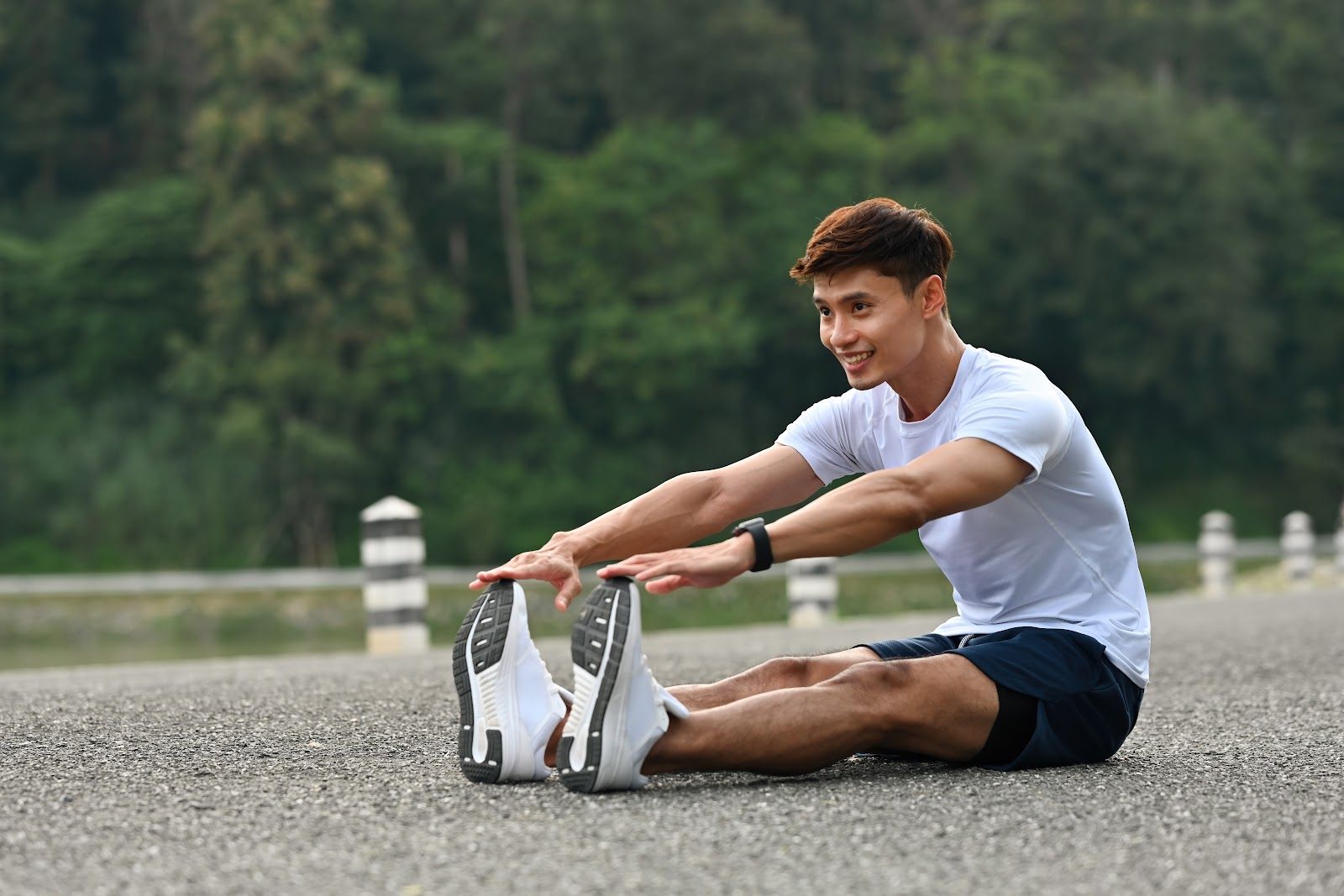 Man sitting on a road stretching forward, wearing a white shirt and black shorts, with trees in the background.