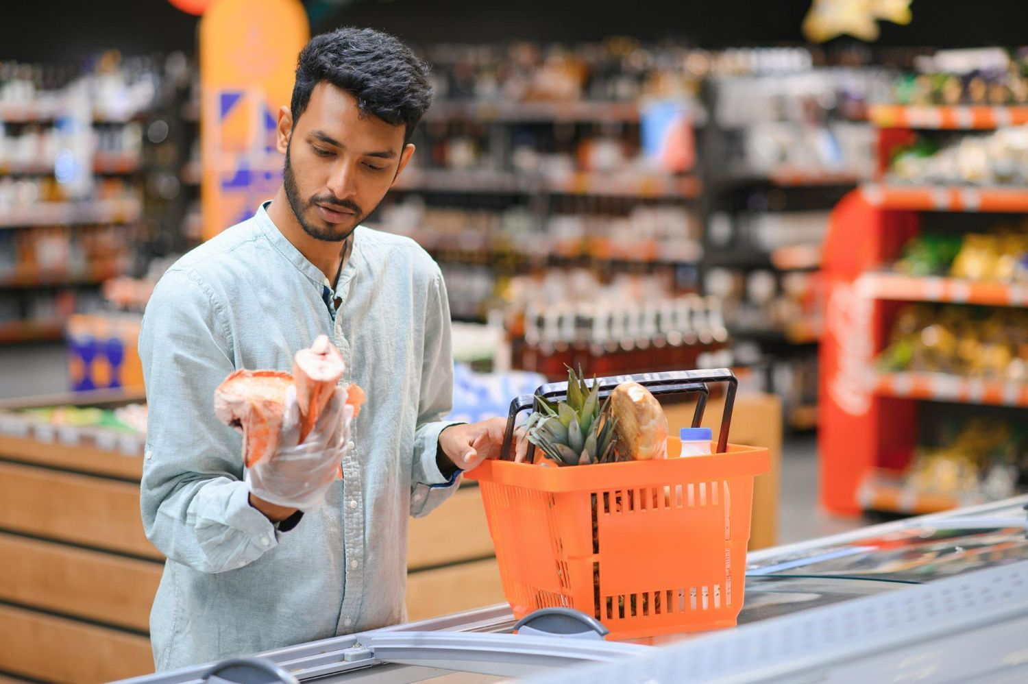 A person wearing a glove holds a piece of food while standing at a grocery store, next to an orange shopping basket.