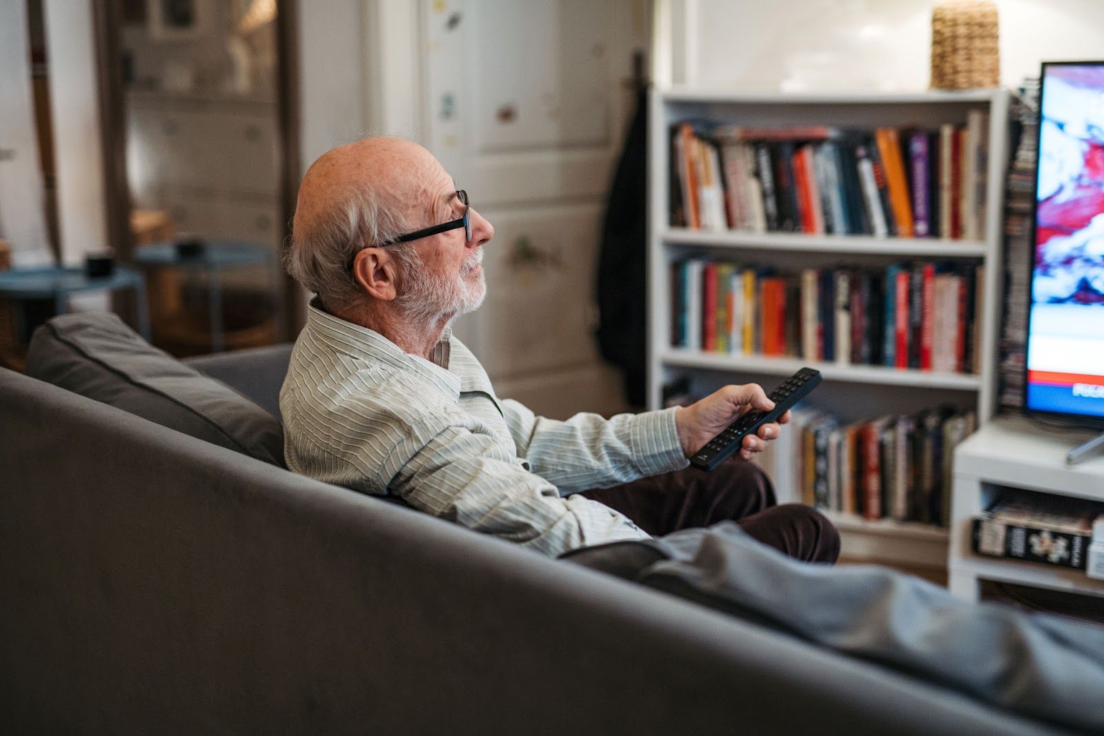 Older man with glasses using a remote on a couch, watching TV in a living room