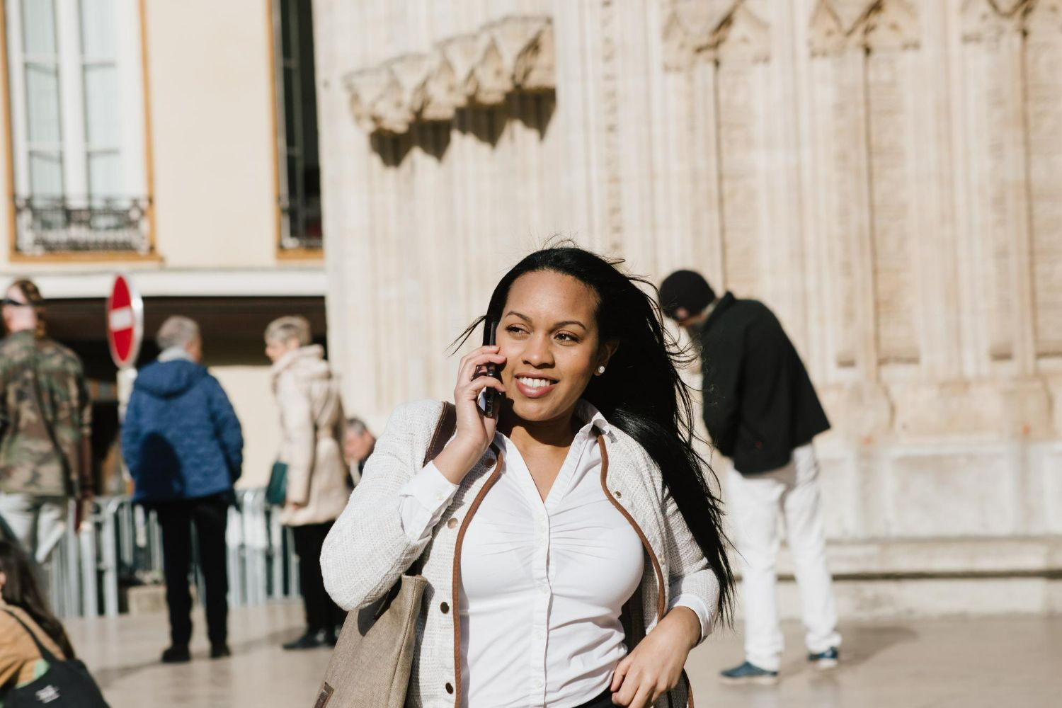 A woman talking on a smartphone while walking outdoors in front of a stone building with people in the background.