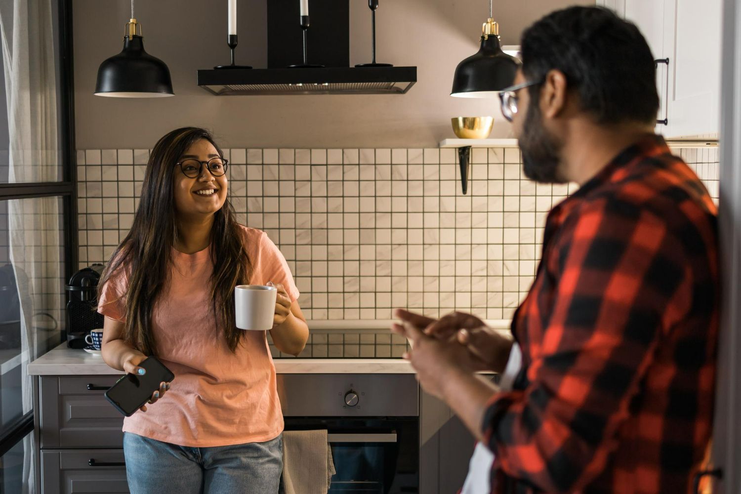 A person holding a coffee mug and phone talks to another person wearing a red flannel shirt in a kitchen.