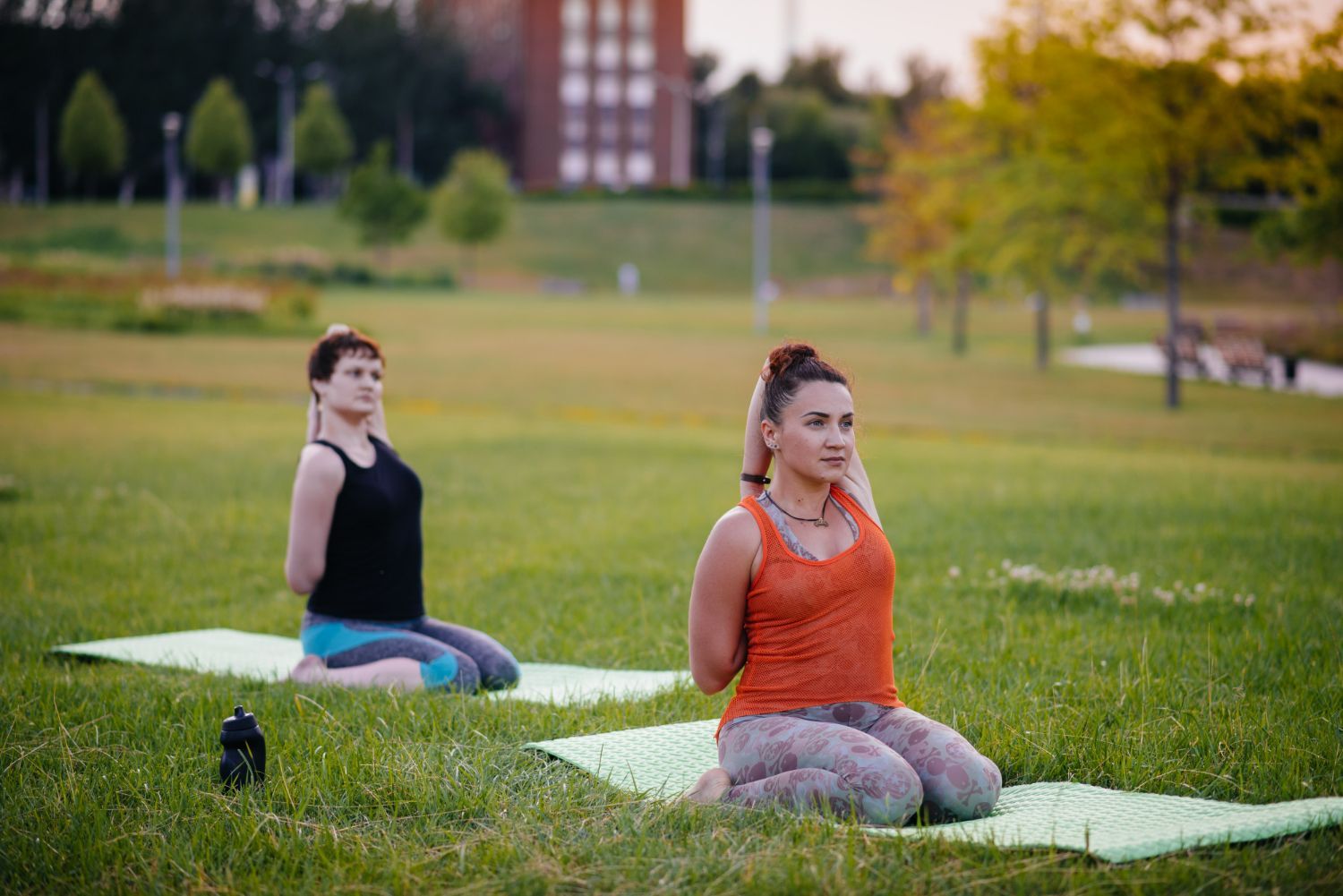 Two people practicing yoga outdoors on green mats in a park, sitting with their arms folded behind their backs.