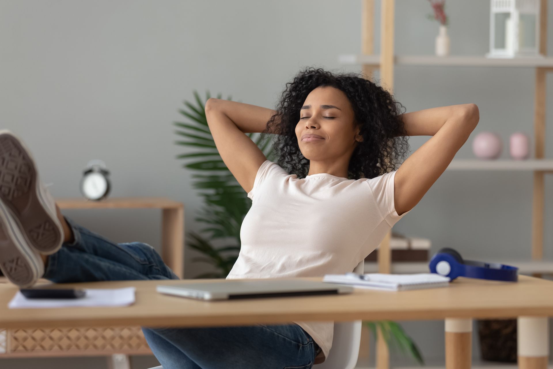 A woman is sitting at a desk with her legs crossed and her hands behind her head.