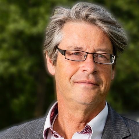 Een man met een bril, grijs haar, gekleed in een colbert en wit overhemd, staat buiten te glimlachen.