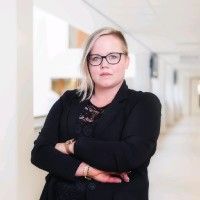 Vrouw met bril en gekruiste armen, gekleed in een zwarte blazer, in een gang.