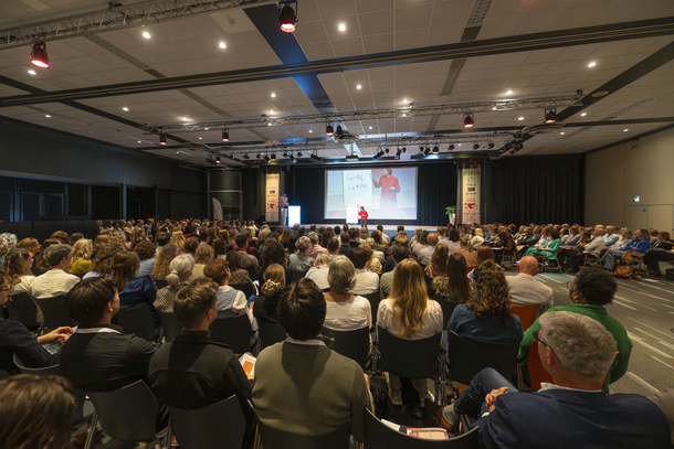 Het publiek op een conferentie luistert naar een spreker op het podium, waar een scherm een ​​afbeelding van een bos toont.