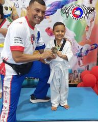 Un hombre con gi blanco y un niño con gi blanco sostienen un trofeo sobre una colchoneta azul en un evento de artes marciales.