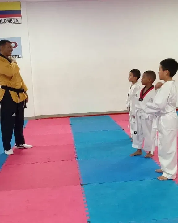 Un instructor con chaqueta amarilla se encuentra frente a tres estudiantes con uniformes blancos sobre una colchoneta rosa y azul en un dojo.