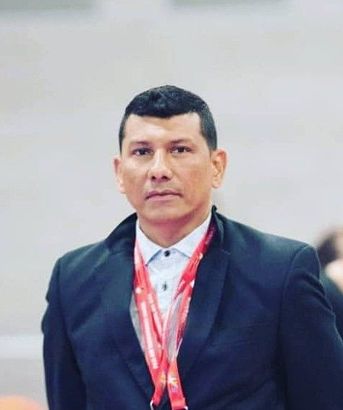 Hombre con traje oscuro y cordón rojo, de pie en el interior de un edificio, con un fondo borroso.
