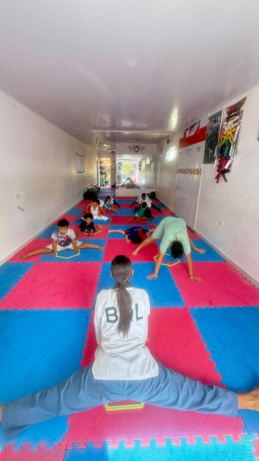 Clase de yoga sobre esterillas rosas y azules en un estudio estrecho, con una persona sentada en primer plano.