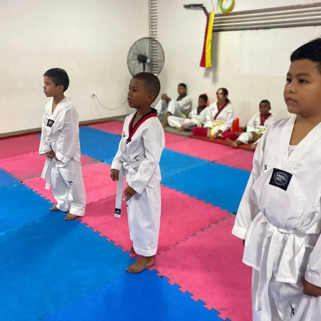 Niños con uniformes blancos de artes marciales de pie en un dojo sobre esterillas rosas y azules.