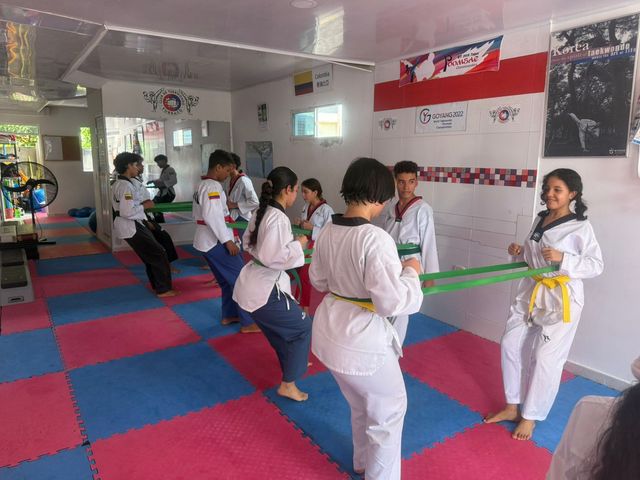 Niños practicando artes marciales en un estudio luminoso con colchonetas rosas y espejos, algunos sosteniendo cinturones de colores.