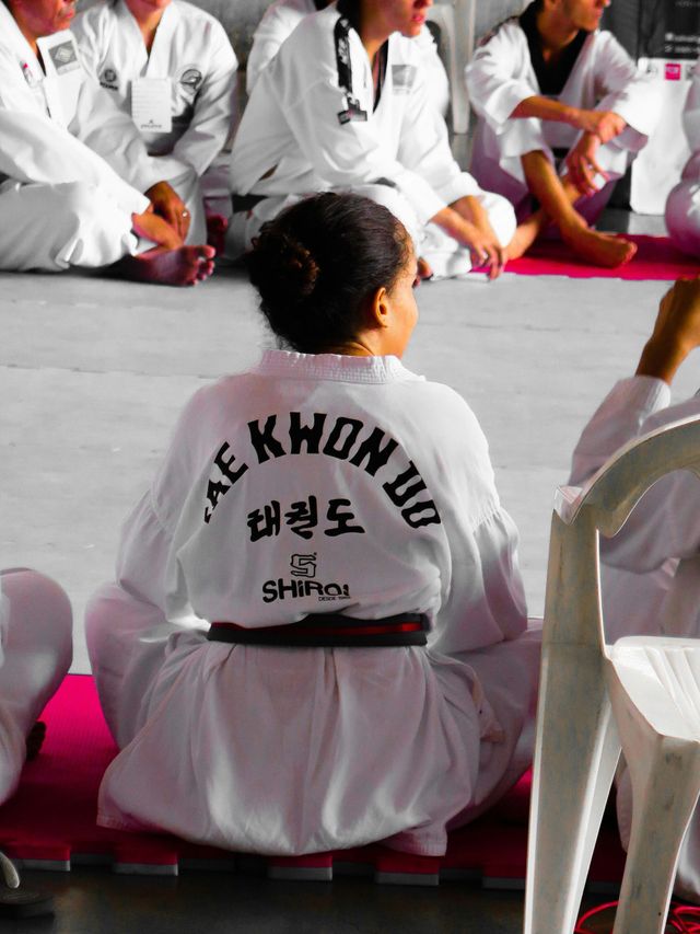 Estudiantes de taekwondo con uniformes blancos sentados en colchonetas, vistos desde atrás durante la clase.