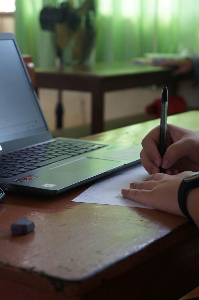 Una persona escribe en un papel junto a un ordenador portátil en un escritorio de madera con una cortina verde al fondo.