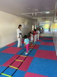 Niños practicando artes marciales sobre colchonetas azules y rojas en un pasillo, guiados por un instructor.