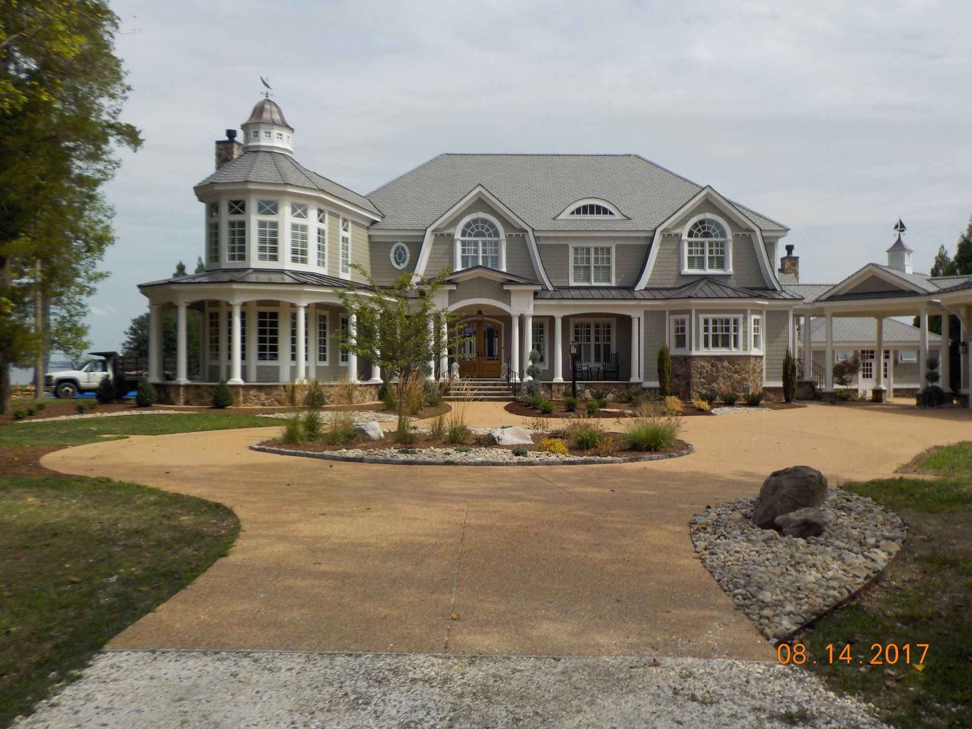 Elegant gray mansion with circular driveway, landscaping, and a turret under a cloudy sky.