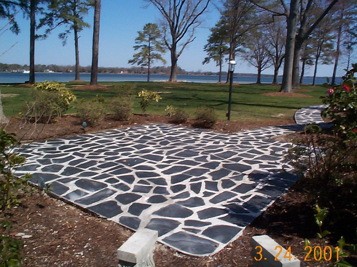 Patio of black irregular stones, edged by concrete, leads to a grassy area by water, trees in the background.