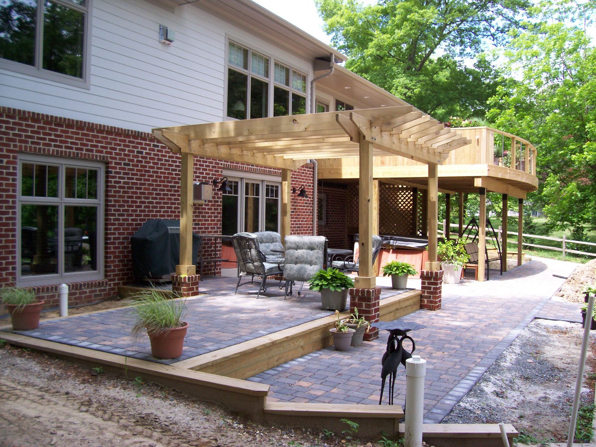 Patio with brick and wood pergola, outdoor furniture, and deck overlooking a wooded area.