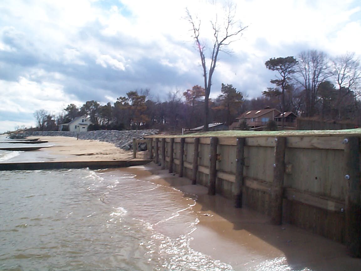 Wooden seawall on a beach, waves lapping against the sand. Cloudy sky, trees in the background.