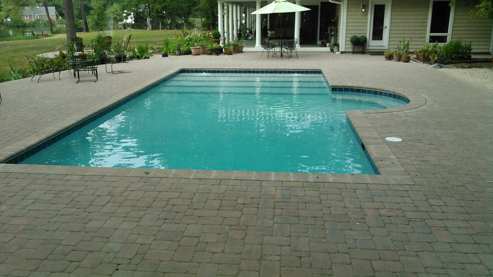 A rectangular swimming pool and attached spa surrounded by brick patio. Green umbrella and table on patio.