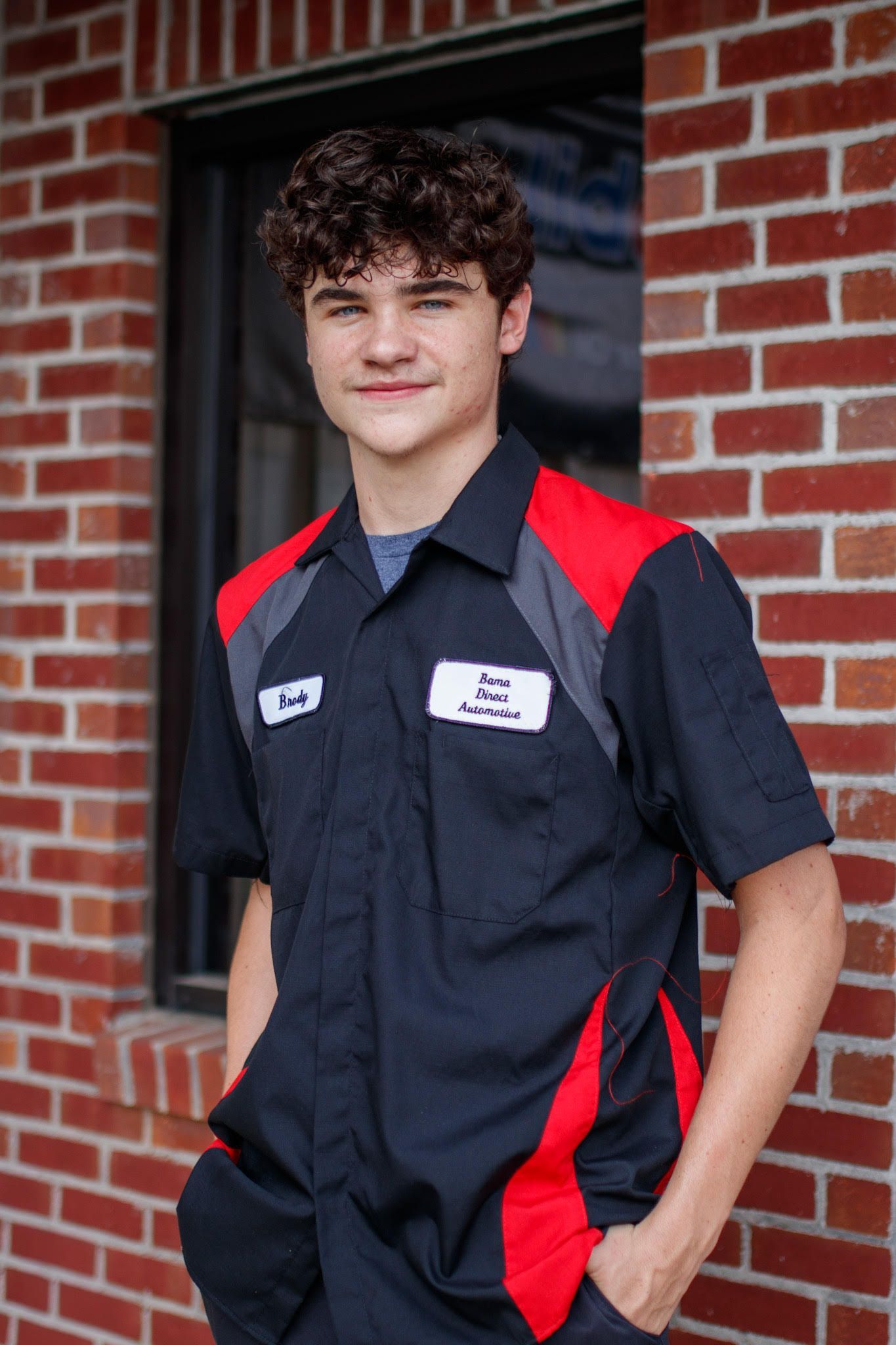 Person in a black and red work shirt standing by a brick wall, hands in pockets, smiling. | Bama Direct Automotive Llc