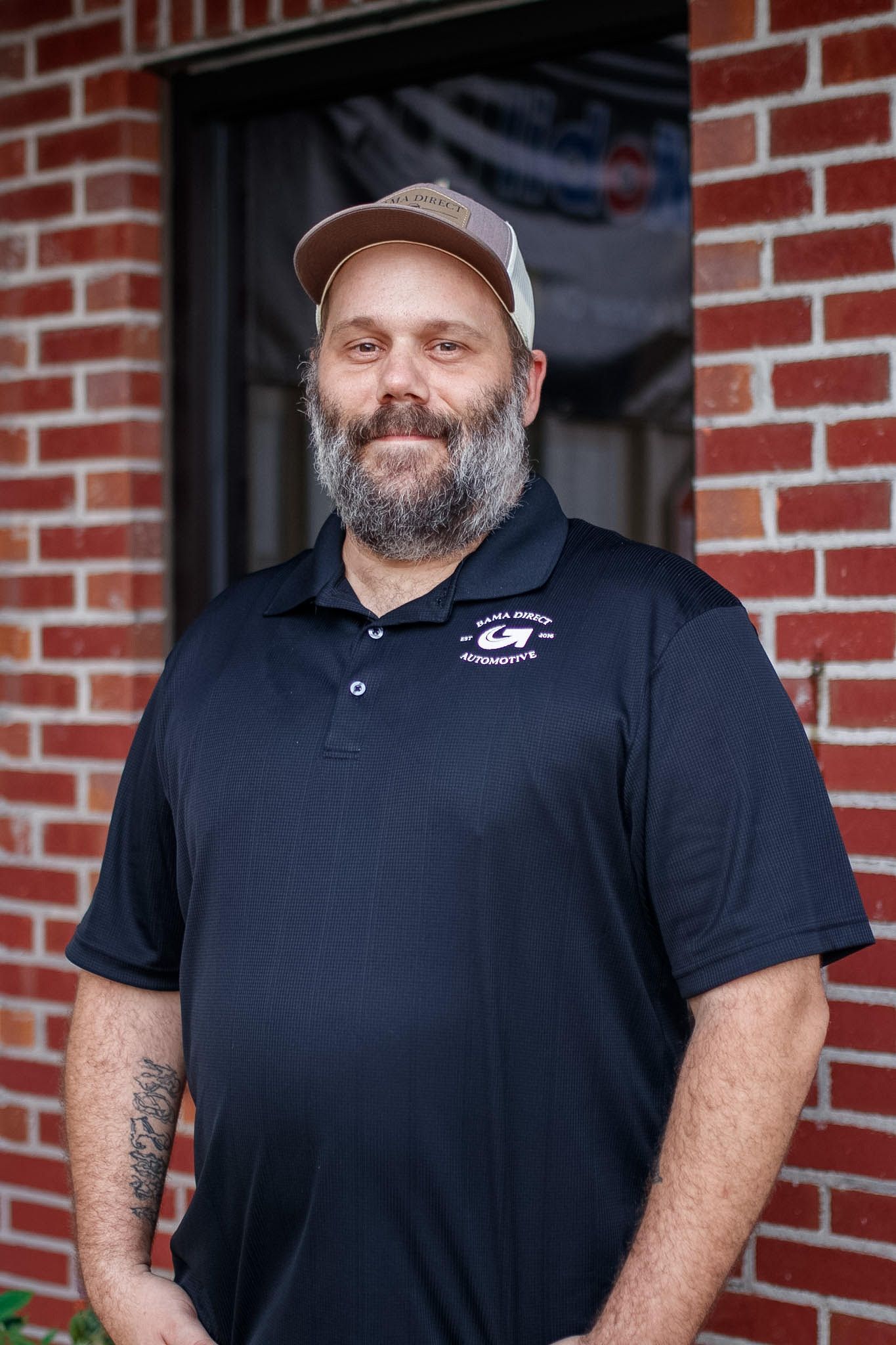 Bearded man in a black polo and tan cap standing in front of a brick wall and window | Bama Direct Automotive Llc