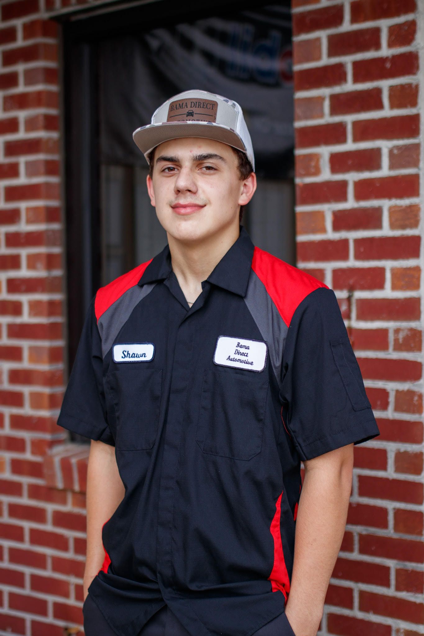 Person in a black and red uniform shirt and cap standing in a doorway by a brick wall. | Bama Direct Automotive Llc