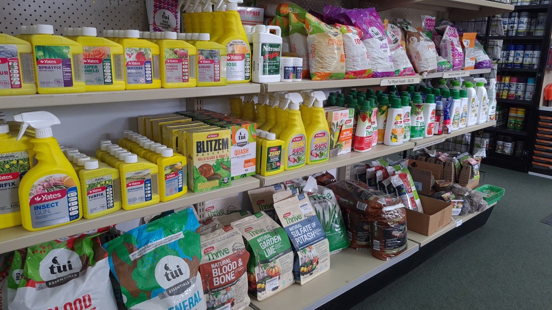 Shelves in a store stocked with yellow cleaning spray bottles, bags of produce, and various household supplies.