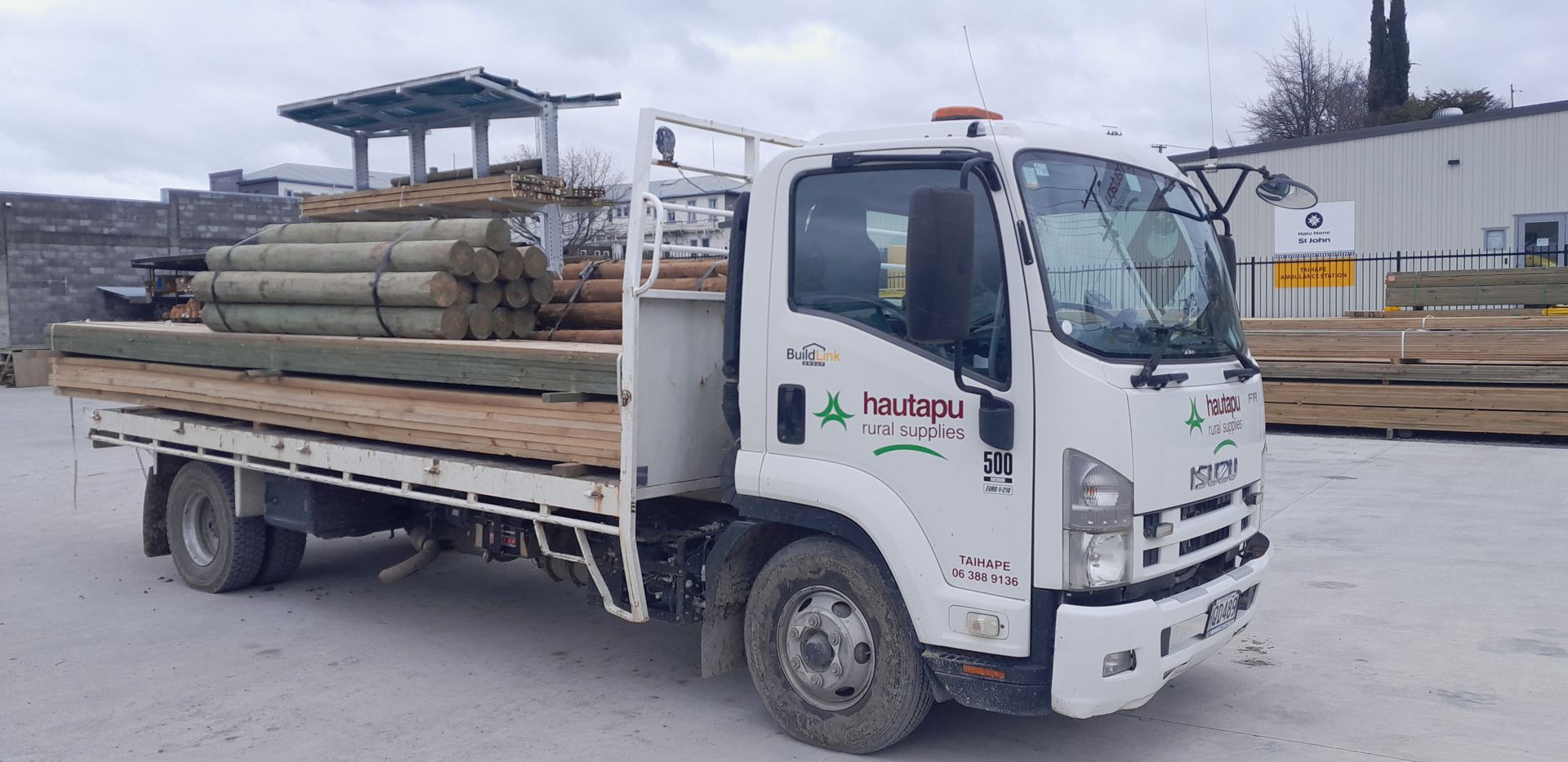 A white Isuzu flatbed truck loaded with stacked timber logs parked on an outdoor lot.