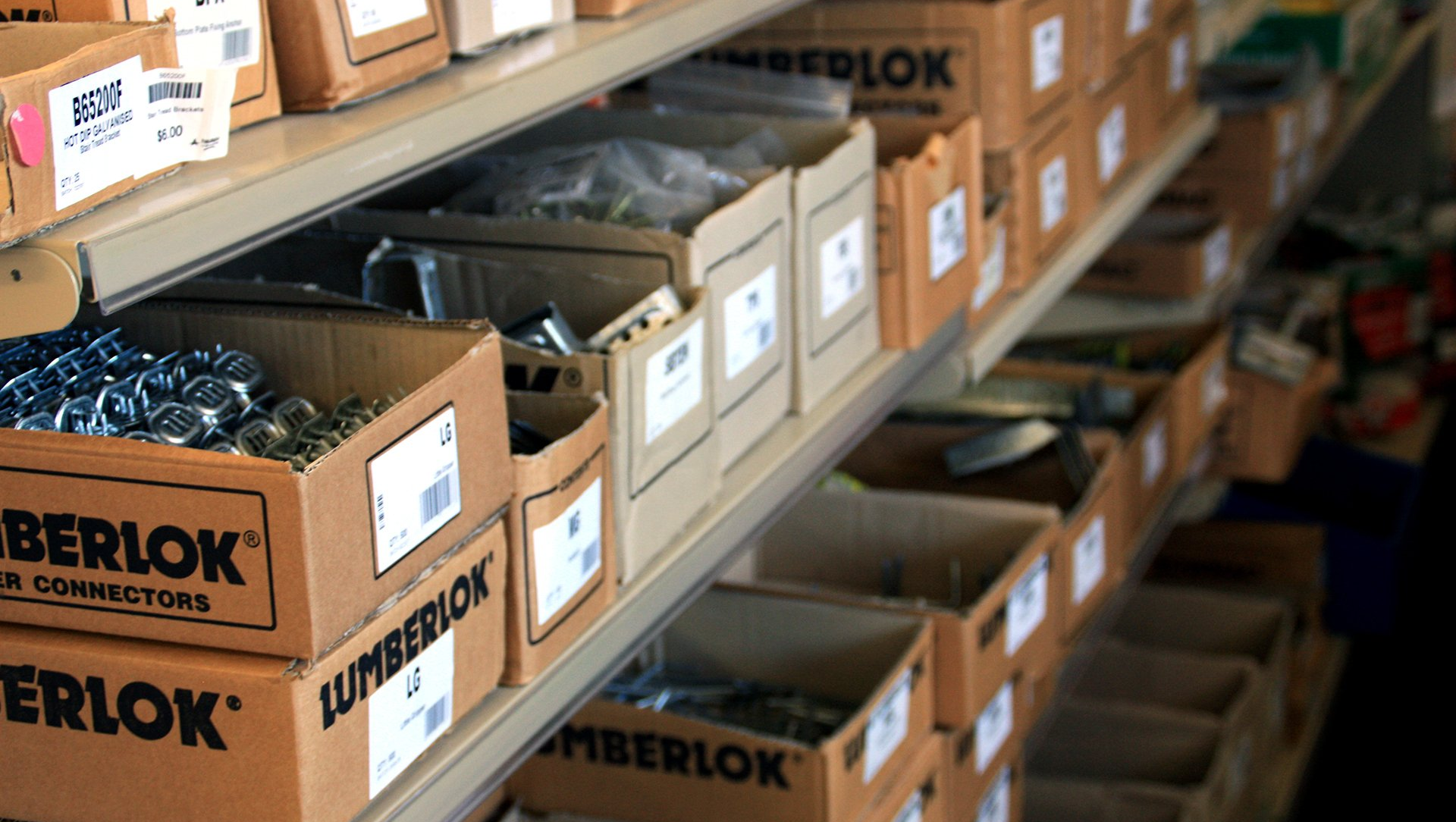 A side view of shelves in a hardware store stocked with labeled cardboard boxes of Lumberlok building connectors.