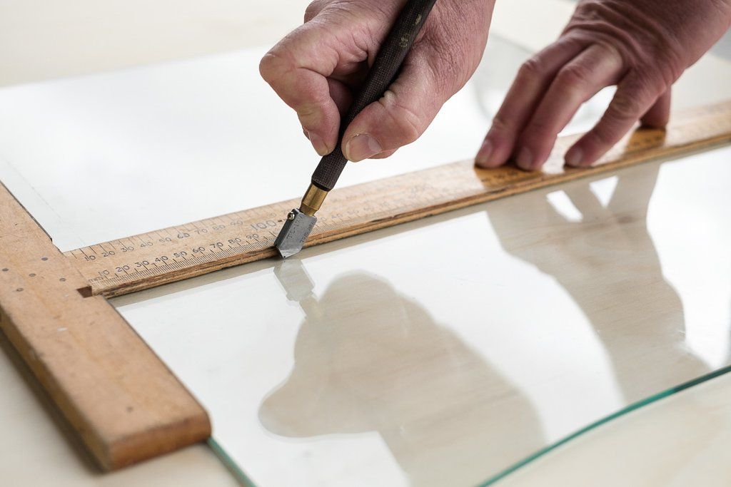 A hand uses a glass cutter along a wooden T-square to score a piece of glass on a workspace.