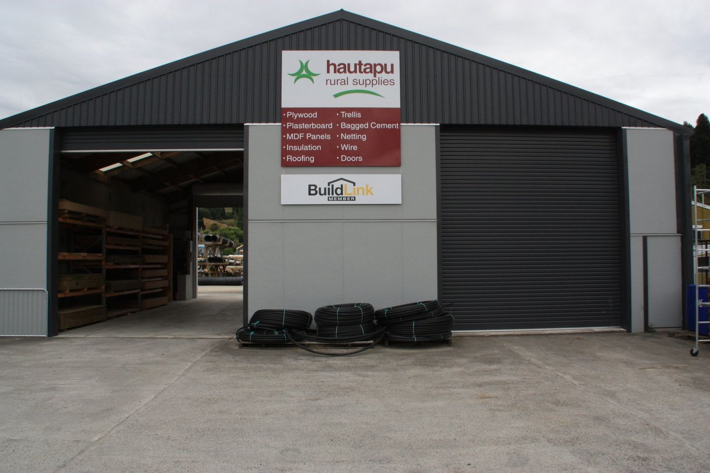 Hautapu Rural Supplies store front with a large metal shed, signage, and black coils of material on the concrete ground.