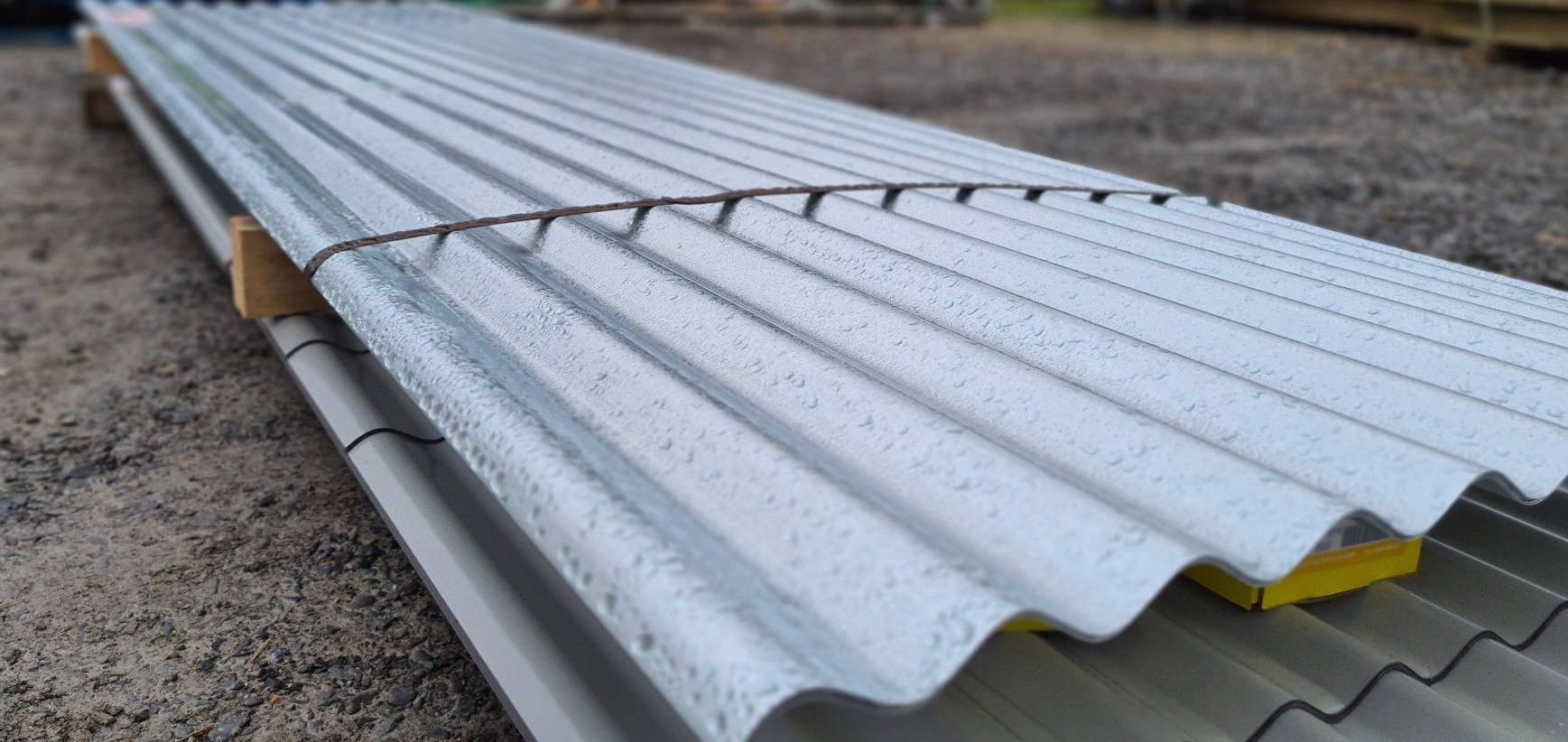 A stack of corrugated metal roofing sheets resting on wooden spacers on a gravel surface.